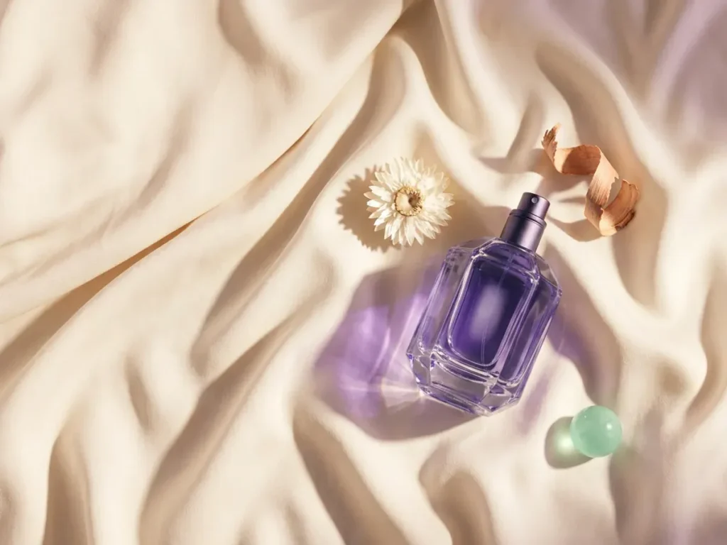 Frasco de perfume facetado violeta sobre seda crema, junto a una flor blanca seca, una viruta de madera y una esfera de cristal verde menta
